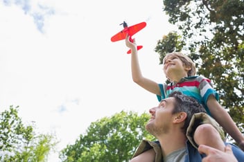Low angle view of a boy with toy aeroplane sitting on father's shoulders at the park